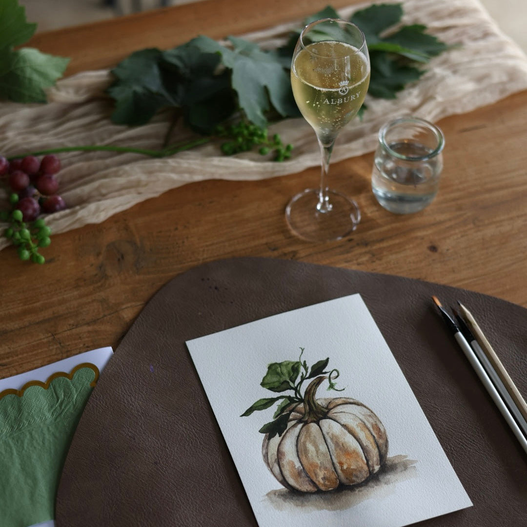 A square art print featuring a watercolor illustration of a light-colored pumpkin with green leaves, displayed on a dark brown leather placemat on a wooden table. The setting includes a glass of white wine, a small glass of water, and grapevine leaves in the background.
