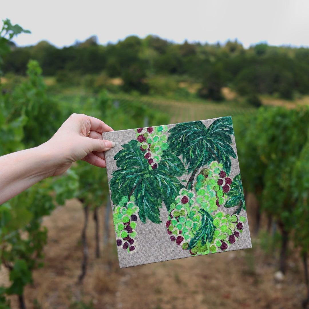 A hand holds a square canvas painting depicting green grapevines and clusters of green and purple grapes against a blurred vineyard background.