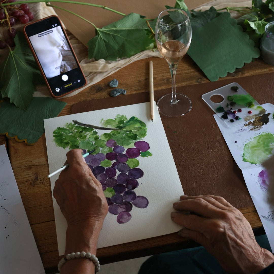 A person's hands painting a watercolor of purple grapes on a white sheet of paper at a wooden table, surrounded by grape leaves, a wine glass, and an artist's palette.