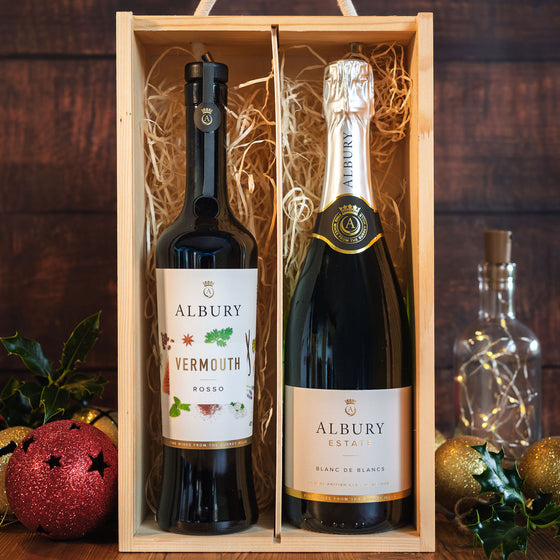 A wooden gift box containing two Albury Estate bottles: a dark red Vermouth Rosso and a sparkling white Blanche de Blanc. The bottles are nestled in natural straw filler, set against a dark wood background with festive holiday decorations including a red ornament, holly, and a glass bottle with fairy lights.