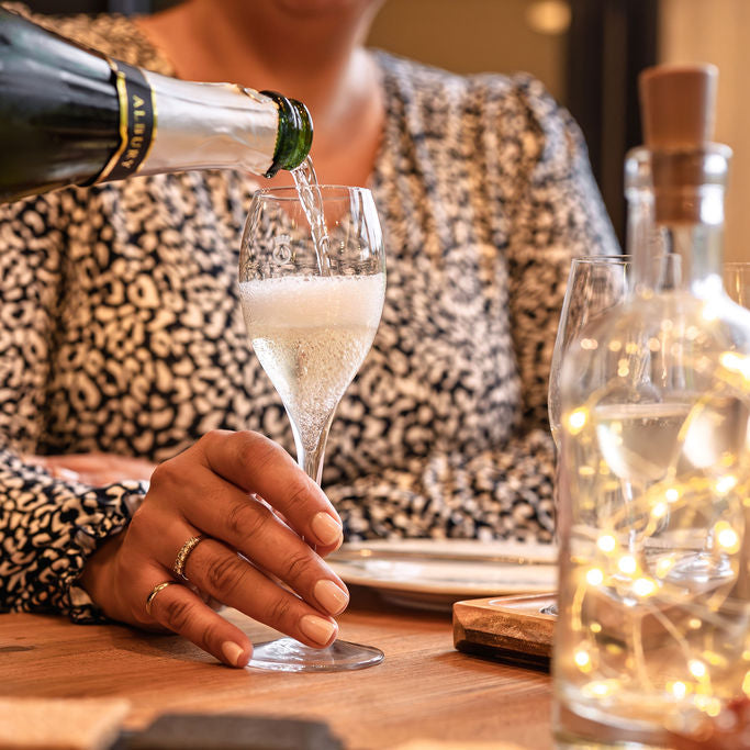 Person pouring champagne into a glass on a table with decorative lights.