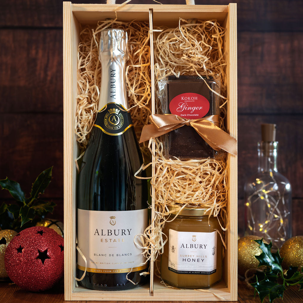 A wooden gift box filled with shredded straw contains a bottle of Albury Estate sparkling wine, a jar of Albury Honey, and artisan chocolates tied with a gold ribbon. The box is set on a dark wooden surface with festive ornaments and a glass bottle in the background.