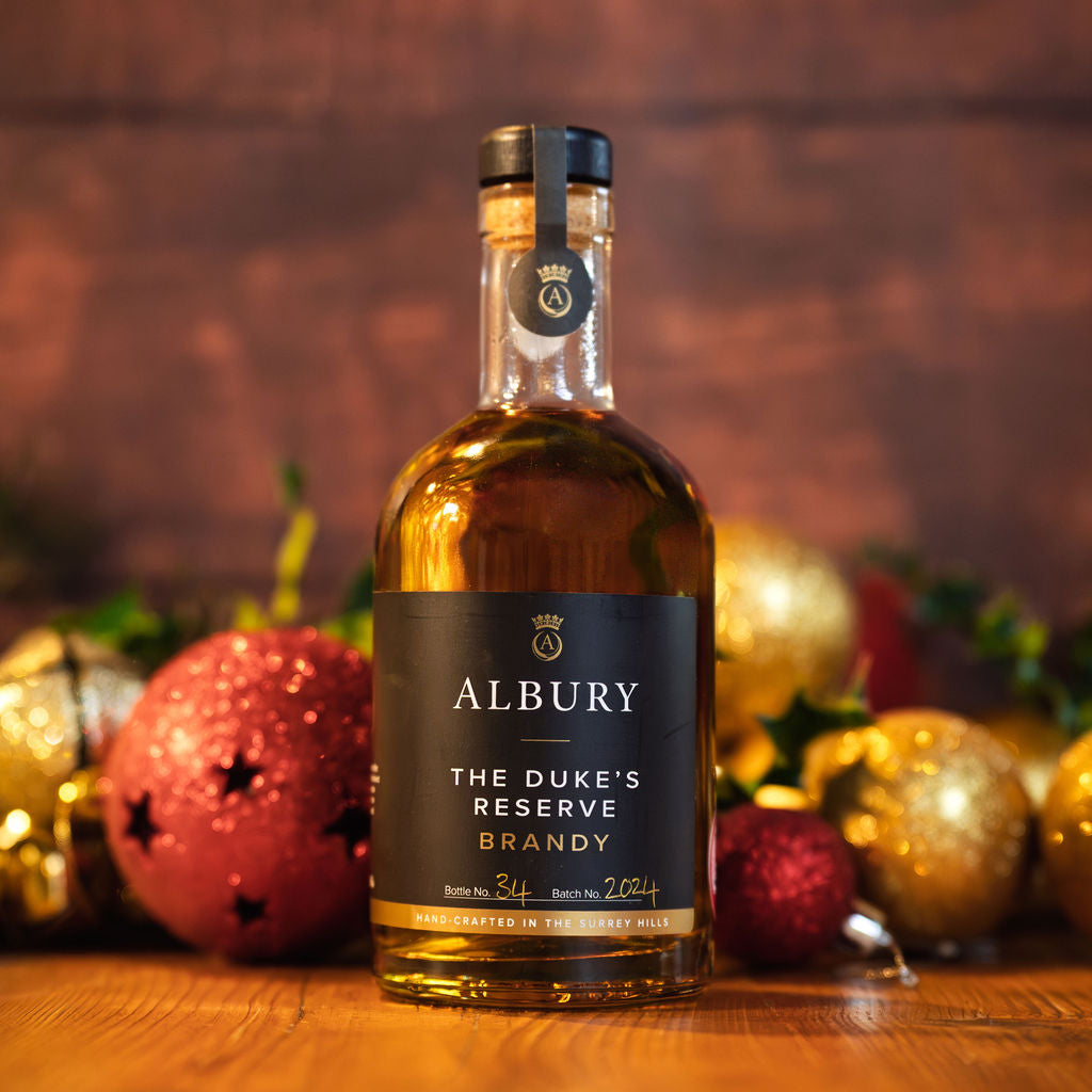 A bottle of Albury The Duke's Reserve Brandy stands on a wooden surface with festive red and gold ornaments blurred in the background against a warm brown backdrop.