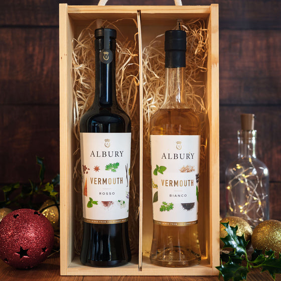 A wooden gift box containing two Albury Vermouth bottles, one dark red and one pale white, displayed on a dark wooden surface with holiday decorations.