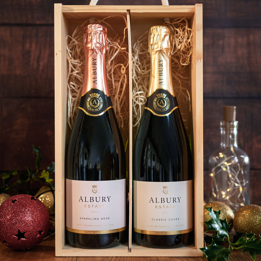 Two Albury Estate sparkling wines in a light wood gift box with shredded paper, set against a dark wood background with festive decorations.