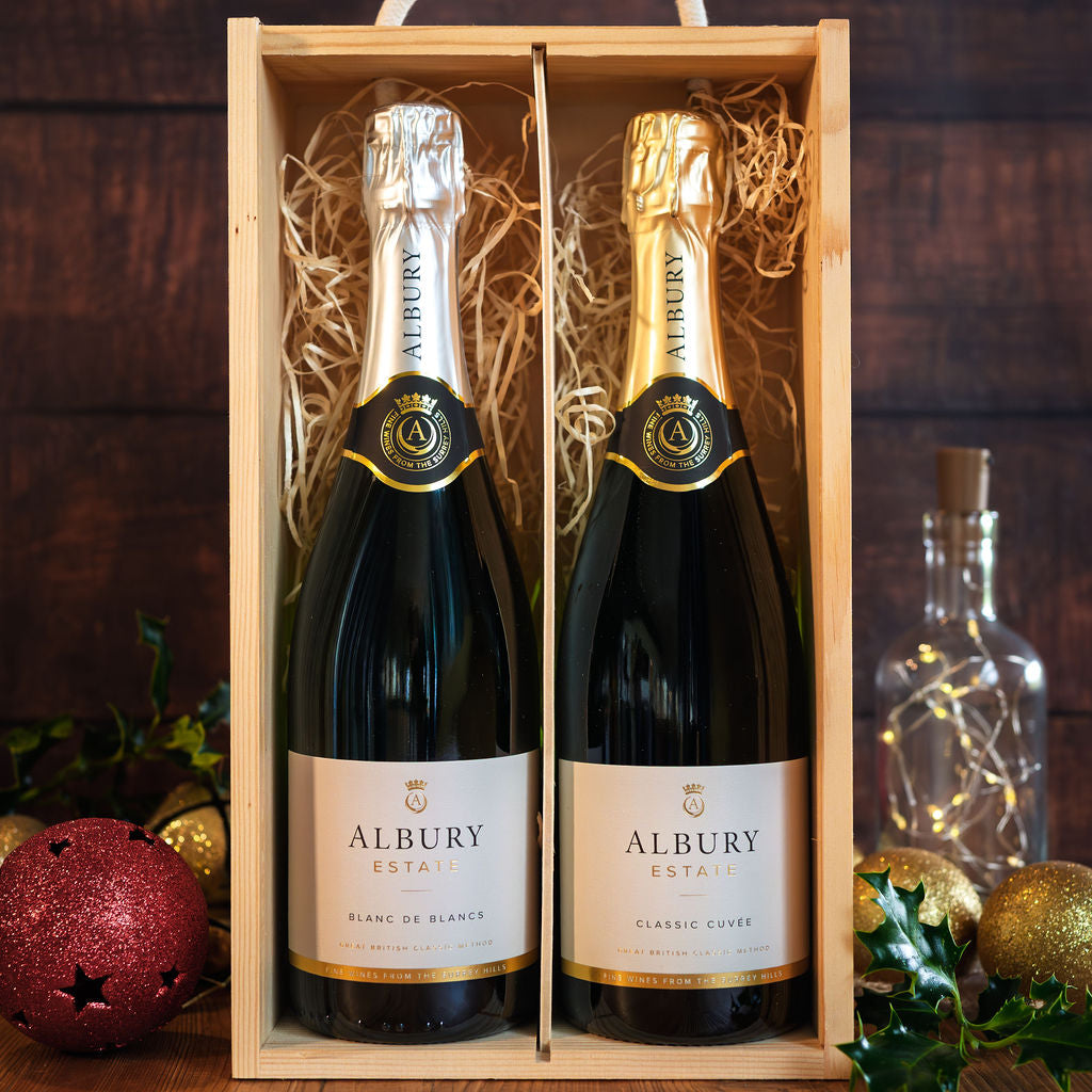 Two Albury Estate sparkling wine bottles presented in a light wood gift box with shredded paper filler, set against a dark wood background with holiday decorations.