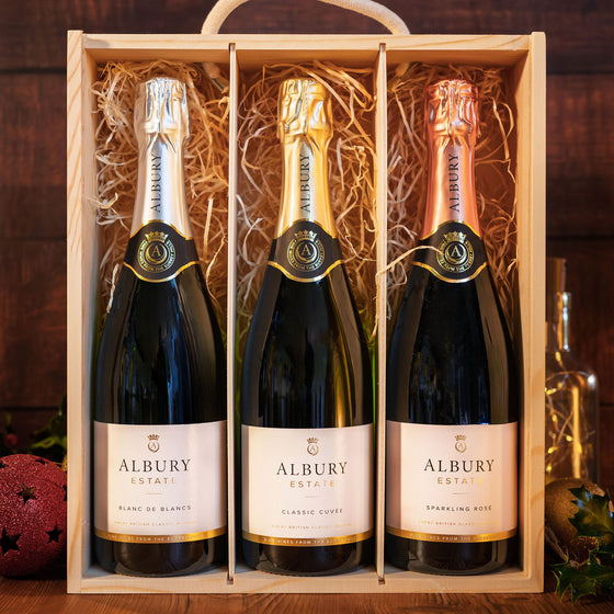 Three Albury Estate sparkling wine bottles in a light wood gift box with shredded paper filler, set against a dark wooden background.