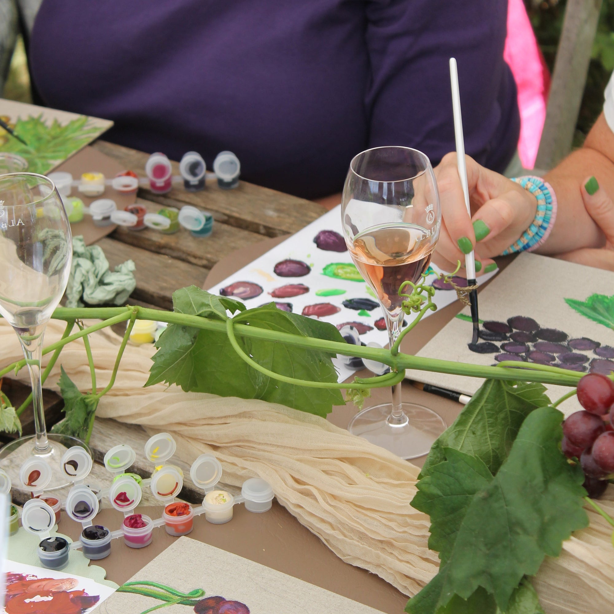 A lifestyle scene of a group painting grapevines outdoors on a wooden table, with wine glasses, small paint pots, and grape illustrations visible.