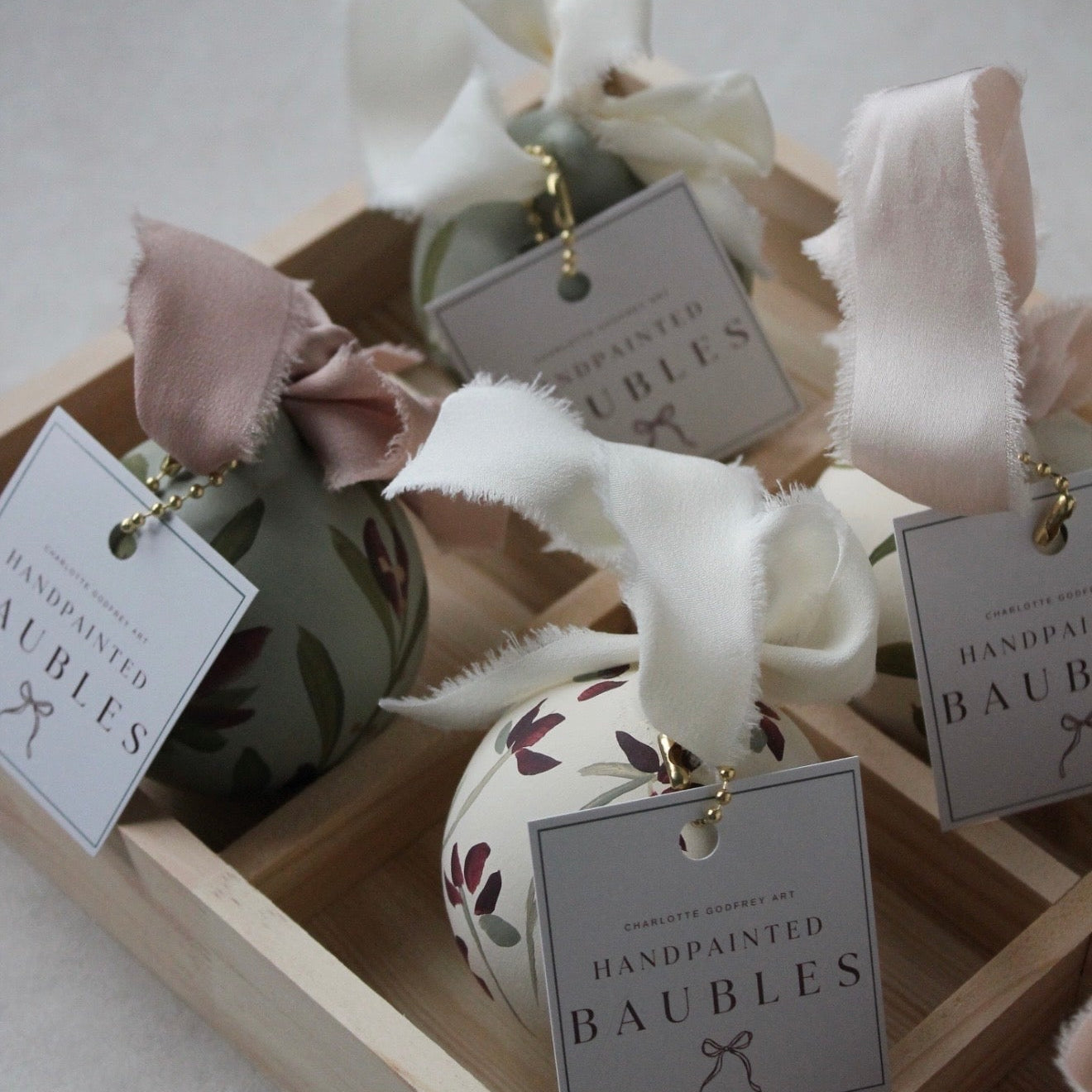 Three hand-painted decorative ornaments displayed in a wooden tray, each with a fabric ribbon bow and a hanging tag that reads 'Handcrafted and Handpainted Baubles.' The ornaments have muted floral patterns in olive green, cream, and blush tones.