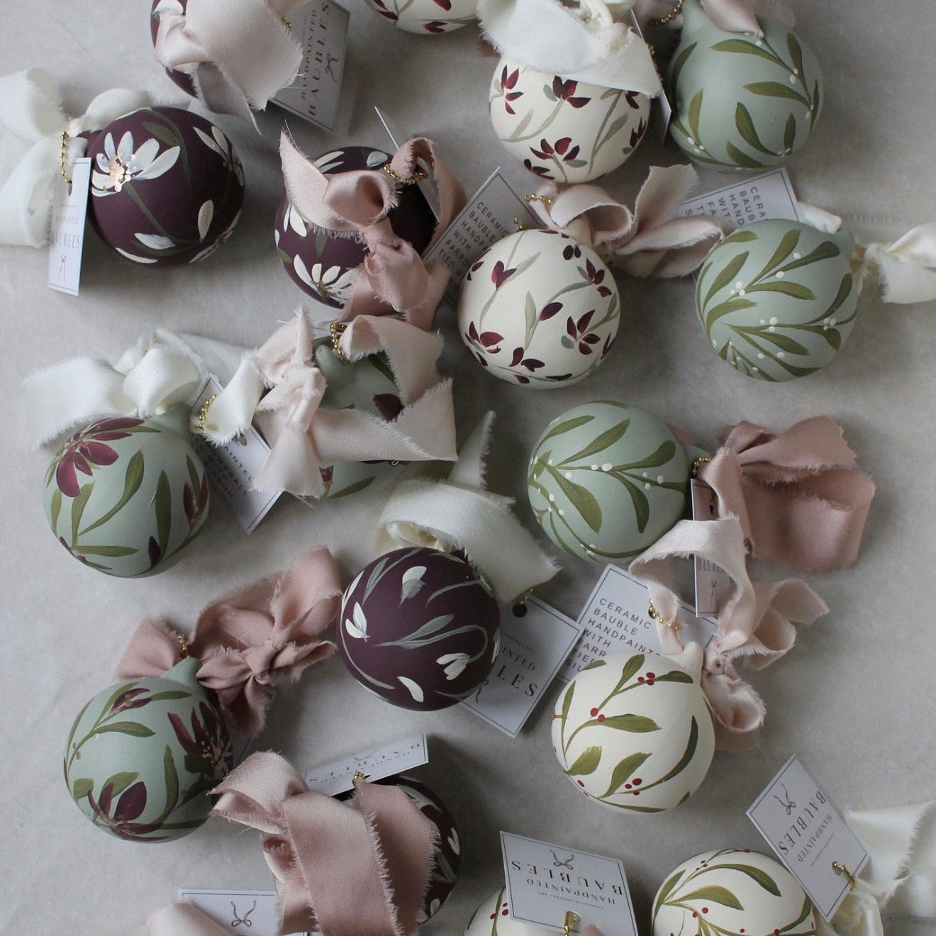 A collection of decorative spherical ornaments with floral and botanical patterns in muted green, burgundy, cream, and blush pink, each tied with a matching ribbon and a small paper tag.