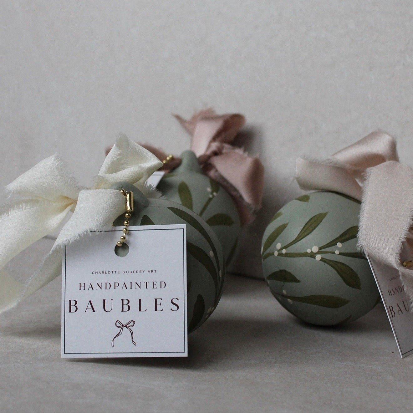 Three handpainted decorative baubles in a muted sage green color with olive leaf patterns, adorned with white and blush pink ribbons and hanging tags that read 'Handpainted Baubles' and 'Charlotte Johnson Art'.