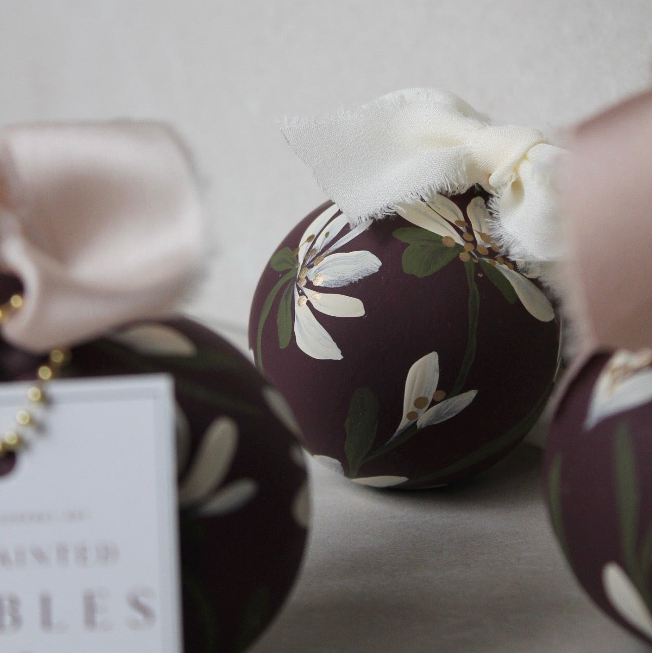 A close-up of a decorative spherical ornament with a dark plum-brown background, white floral print, green leaves, and a white ribbon bow. Part of another matching ornament and a hanging card are visible in the foreground and background.
