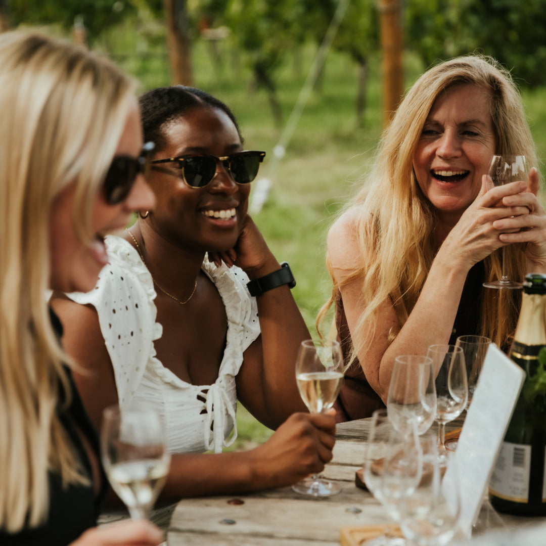 Three women enjoying a wine tasting outdoors with glasses and a bottle on a wooden table.